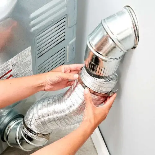 A person’s hands are connecting a flexible, shiny dryer vent hose to a wall duct behind a dryer in a laundry area—an important step for proper airflow and safety, just like regular air duct cleaning Dallas professionals recommend.