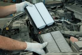 A person wearing white gloves is replacing a rectangular air filter in a car engine compartment, similar to the care shown by air duct cleaning Dallas professionals. They hold a new filter above the old one.