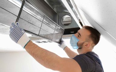 A man wearing a protective mask and gloves is removing or installing an air filter from a ceiling air duct, performing maintenance or cleaning work as part of air duct cleaning Dallas services.